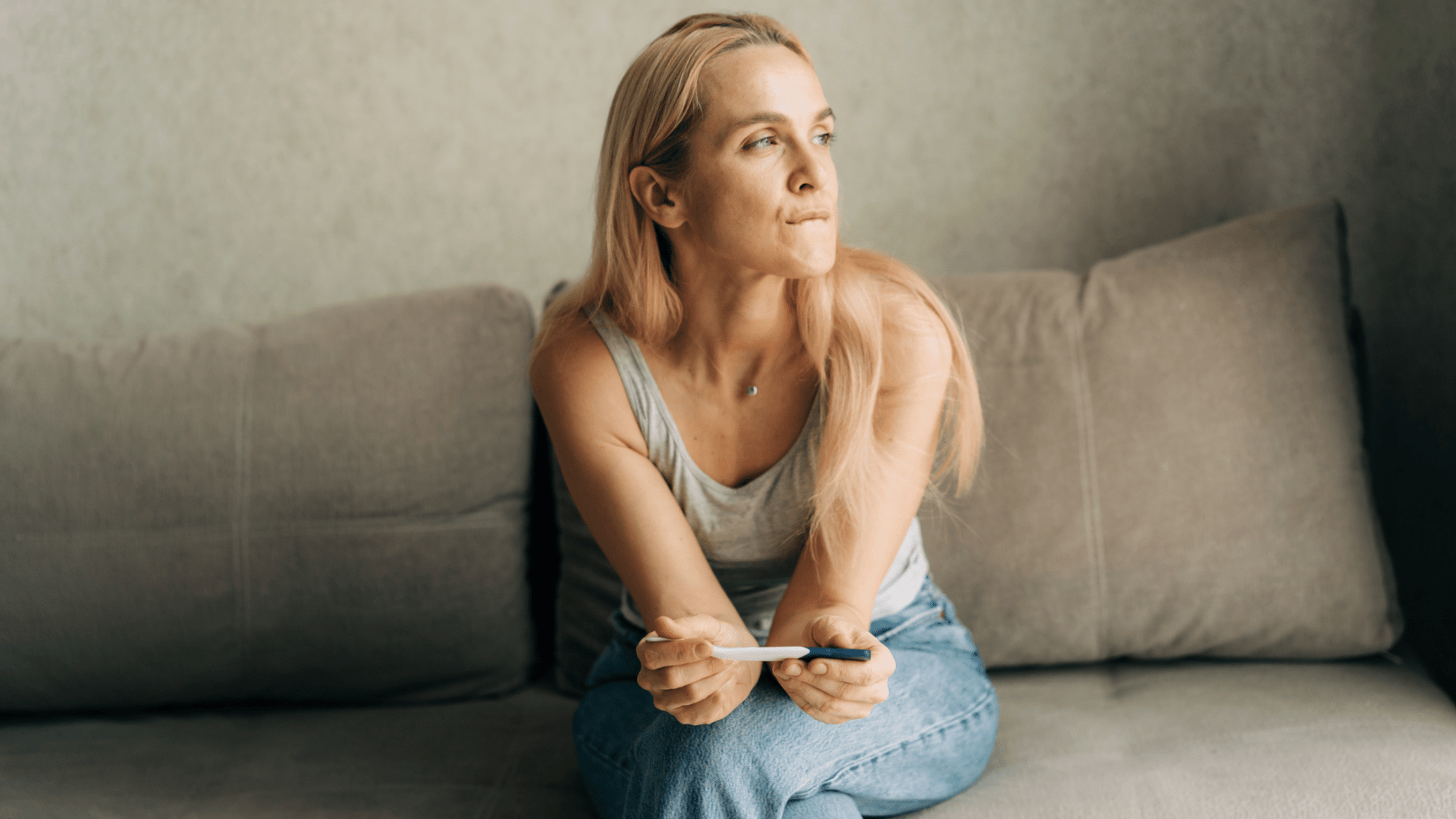 A woman sits thoughtfully on a couch holding a pregnancy test, reflecting on early pregnancy decisions.