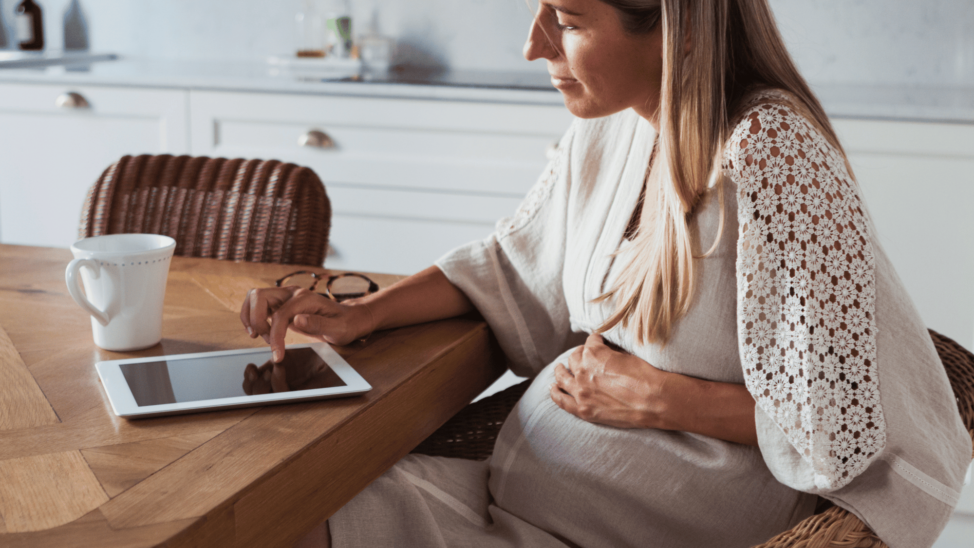 Pregnant woman sitting at a kitchen table using a tablet—engaging in online infant care education at home.