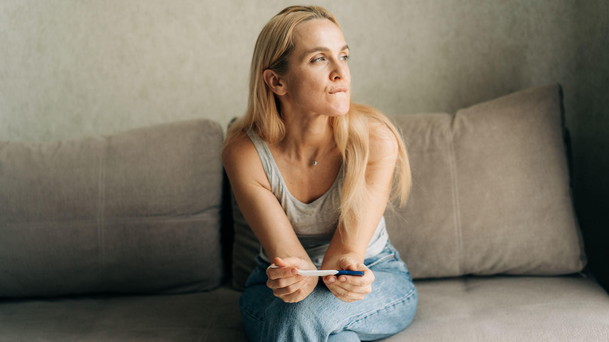 A woman sits thoughtfully on a couch holding a pregnancy test, reflecting on early pregnancy decisions.