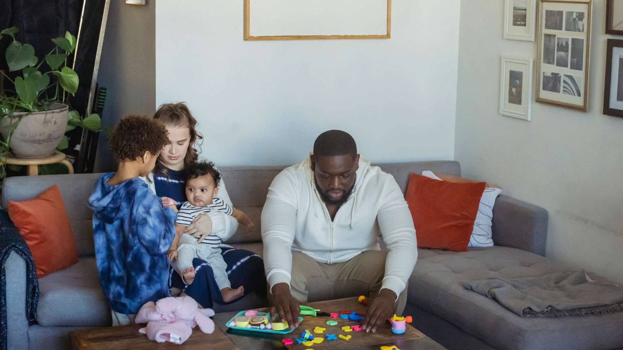 Father playing with colorful developmental toys and building blocks on coffee table while mother holds baby on couch, demonstrating family playtime and age-appropriate toys for toddlers