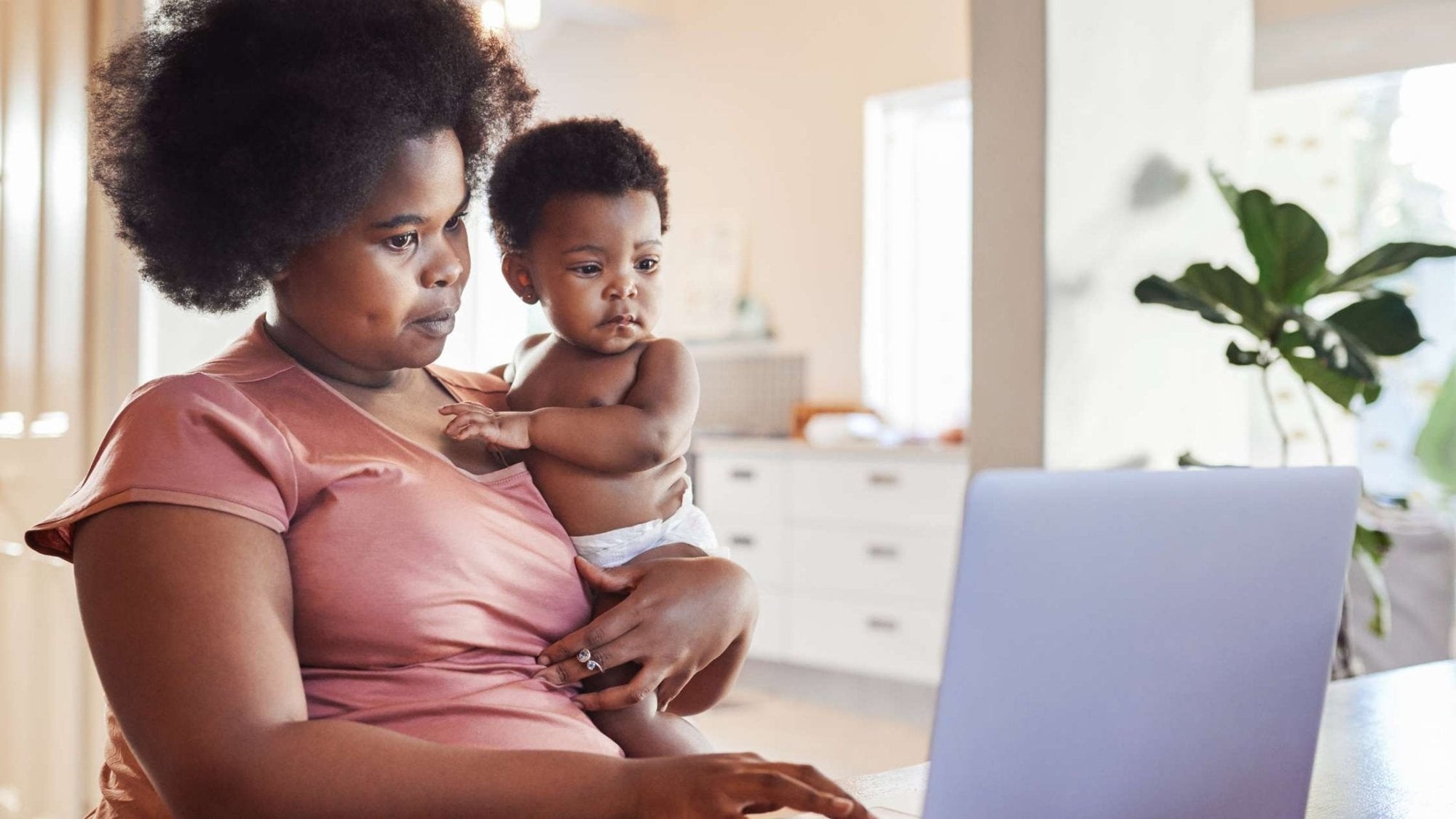 Parent holding baby while working on a laptop, researching feeding cues and starting solids at home