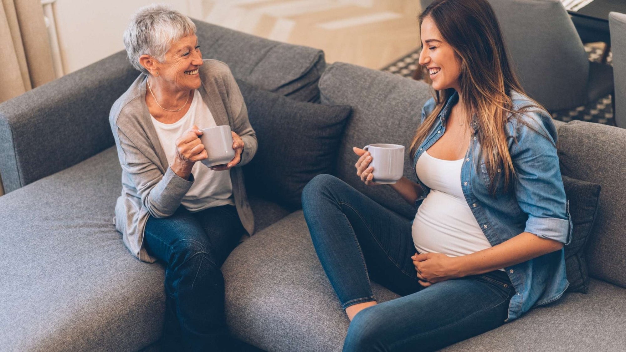 Pregnant woman and older woman share a joyful conversation over coffee on a couch, showing warmth, support, and connection across generations.