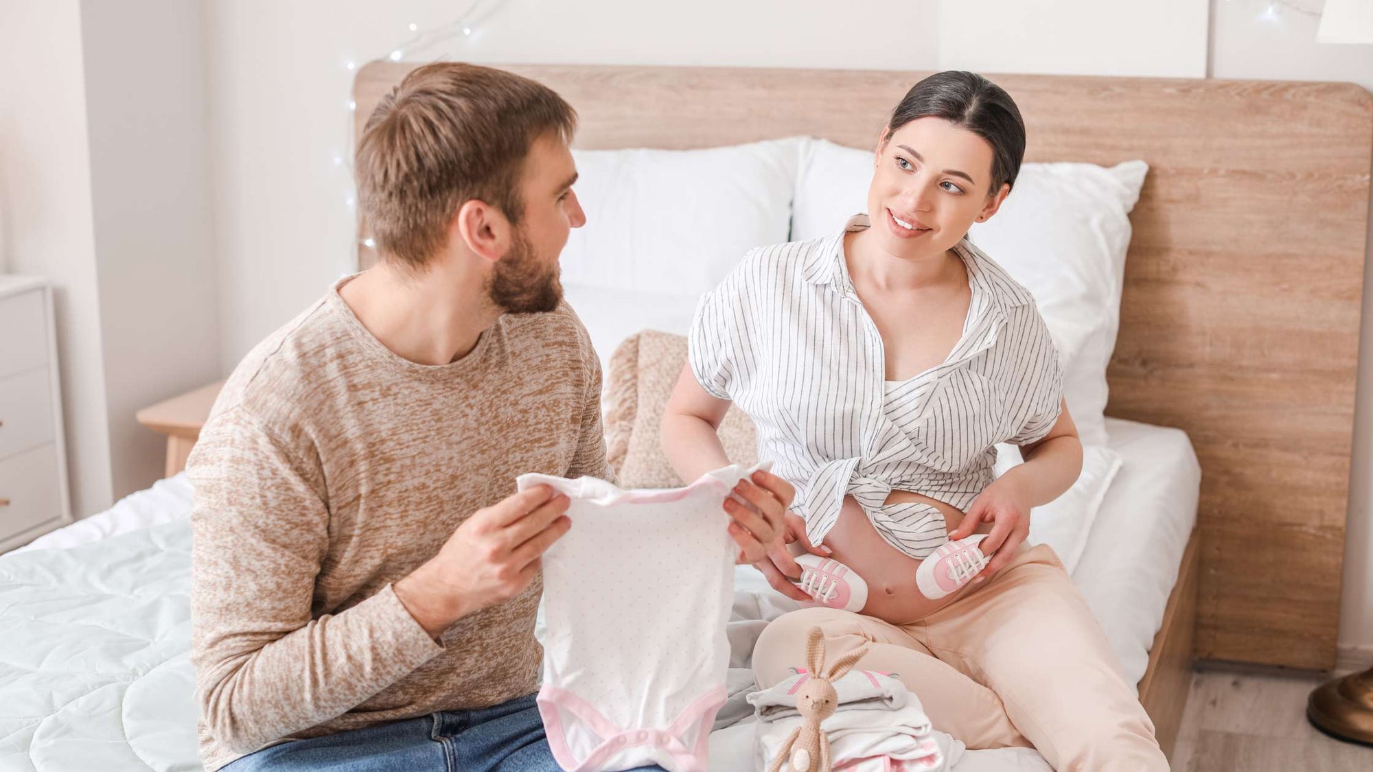  Expecting couple preparing for baby, with mother holding baby booties and father holding a onesie on a bed.